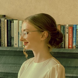 Profile image of the left side of Grace Milusich who is standing before a shelf filled with books. She has long blonde hair that is done in a low bun, and is wearing glasses and a while blouse with a rounded neckline. A long curl has escaped the bun and falls on the right side of her face.